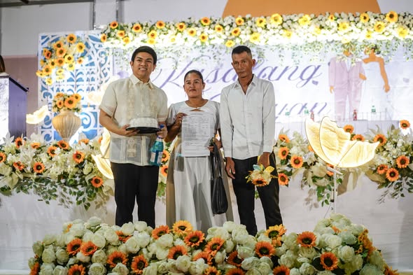 Mga kalahok sa Kasalan ng Bayan 2026 habang hawak ang sertipiko sa entablado na may puti at orange na bulaklak