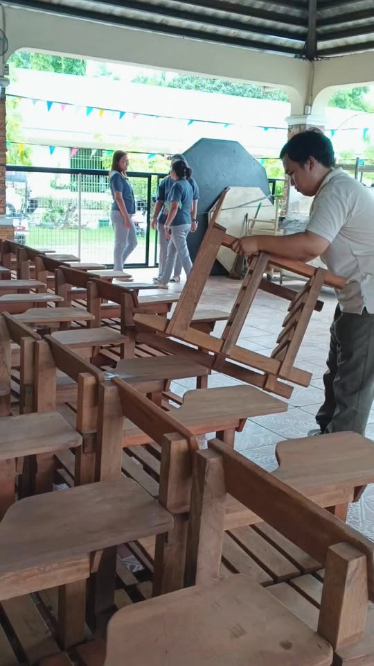 Mga upuang kahoy para sa paaralan na inihahanda sa covered court, habang may lalaking nag-aayos ng isang upuan