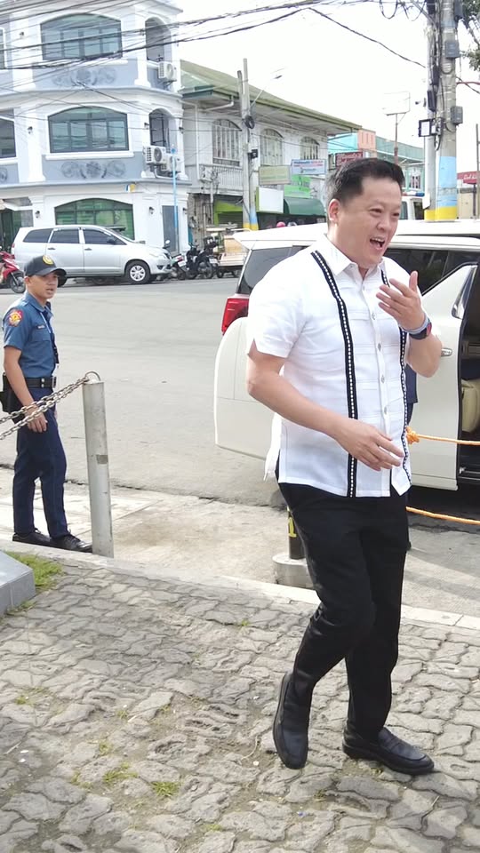 Lalaking naka-barong na naglalakad sa bangketa habang nakangiti, malapit sa puting van at gwardiya sa Naic