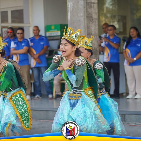 Mga mananayaw na naka-berdeng kasuotan at gintong korona habang nagtatanghal sa harap ng gusali