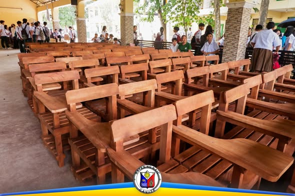 Mga bagong wooden armchairs na nakahanay sa covered hall ng paaralan, may mga estudyanteng nasa likuran
