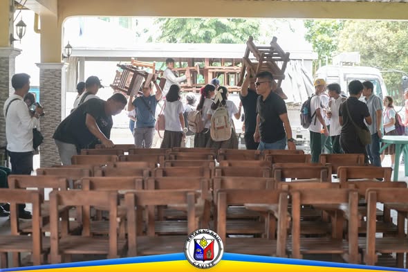 Mga tao na nagbubuhat ng armchairs sa covered area habang inihahanda ang pamamahagi sa mga paaralan sa Naic