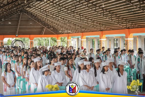 Mga nagtapos ng ALS na naka-puting toga at mortarboard habang nakatayo sa covered hall ng San Roque Elementary School