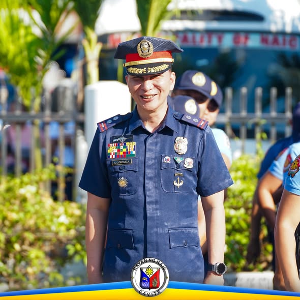 Naka-unipormeng opisyal na nakangiti sa harap ng gusali ng Municipality of Naic habang may flag raising ceremony