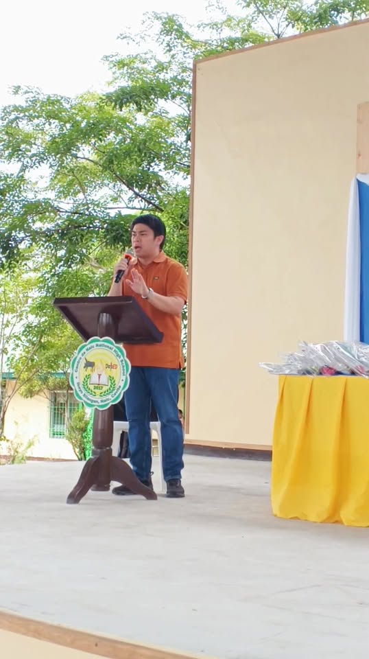 Nagsasalita sa mikropono ang isang opisyal sa podium sa entablado ng Brigada Eskwela sa Centro de Naic NHS