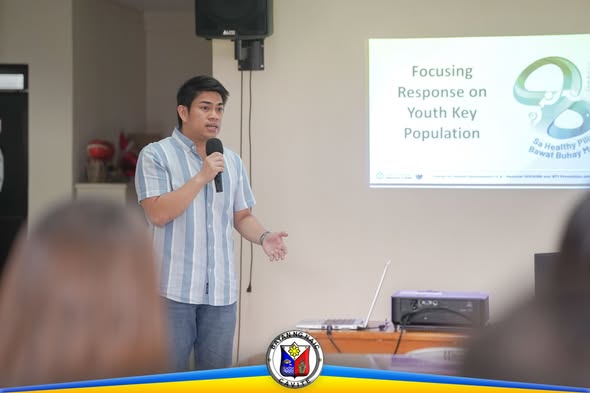 Isang lalaking nagsasalita sa mikropono sa harap ng screen na may presentasyon tungkol sa youth key population