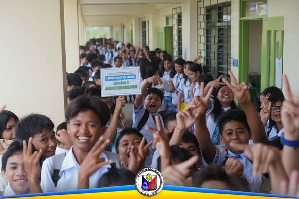 Mga estudyante sa pasilyo ng paaralan, nakangiti at nagpapakuha ng larawan habang may hawak na karatulang "Line starts here"