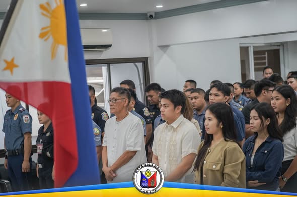 Mga tao at uniformed na kawani na nakatayo habang nasa flag raising ceremony sa Naic Municipal Hall