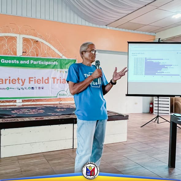Isang lalaki na nagsasalita sa mikropono sa Varietal Field Trial, katabi ang projector screen sa Malainen Luma, Naic