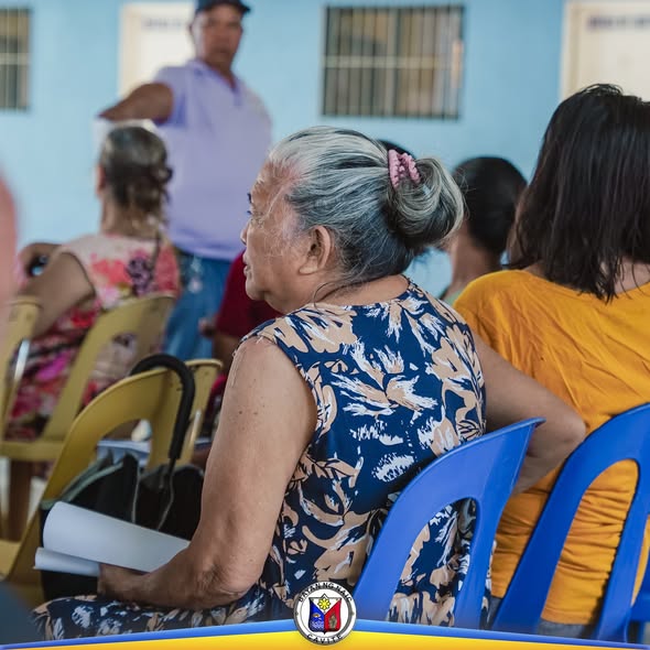 Matandang babae na nakaupo sa covered court habang may nagsasalita sa harap sa pamamahagi ng social pension