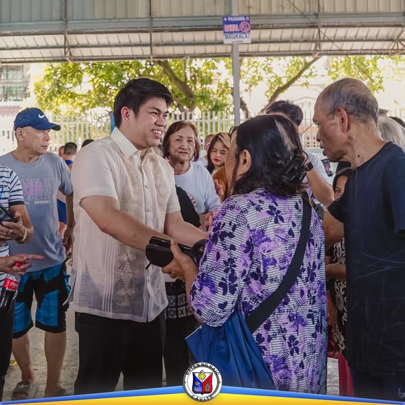 Mga tao sa covered court habang nakikipagkamay ang isang lalaking naka-barong sa isang ginang sa pamamahagi ng pensyon