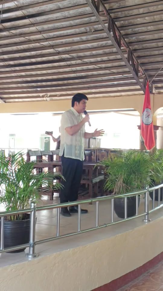 Isang opisyal na nagsasalita sa mikropono sa entablado ng covered hall habang nagaganap ang Brigada Eskwela