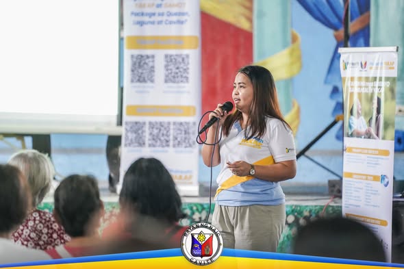 Babaeng nagsasalita sa mikropono sa harap ng mga nakaupong tagapakinig at banner ng PhilHealth YAKAP sa covered court