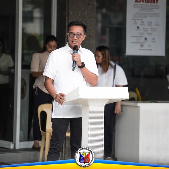 Lalaking nagsasalita sa mikropono sa podium sa harap ng Naic Municipal Hall habang nakikinig ang mga kawani