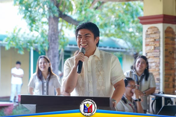 Litrato: Lalaking nagsasalita sa mikropono sa flag ceremony sa Naic INHS, may mga kasama sa likuran