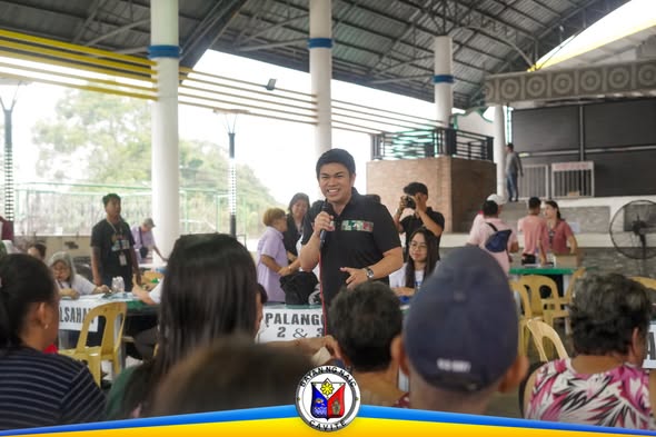 Nagsasalita sa mikropono ang isang lalaki sa harap ng mga senior citizen sa National Social Pension Distribution sa Naic