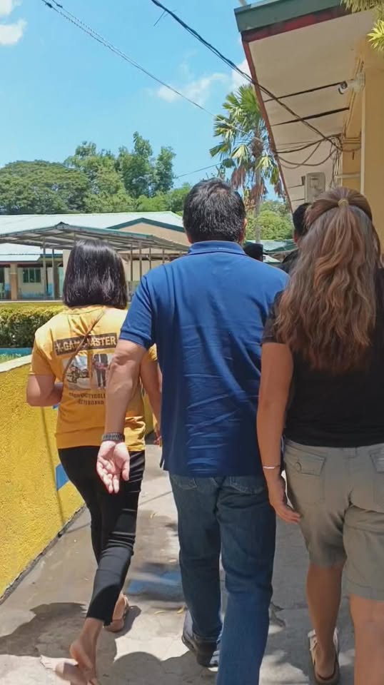 Mga tao naglalakad sa pathway ng paaralan papunta sa gusali habang may dalang school chairs sa Timalan