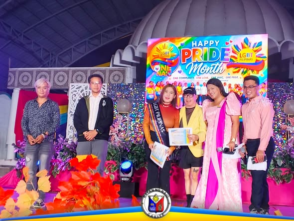 Mga tao sa entablado sa SAGAYLA 2026 sa Naic Town Plaza, may hawak na sertipiko sa ilalim ng Happy Pride Month backdrop