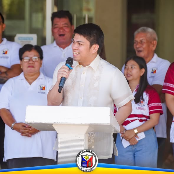 Lalaking nagsasalita sa mikropono sa podium habang nakikinig ang mga kawani sa flag raising ceremony sa Naic