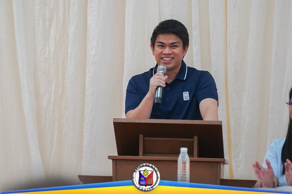 Isang lalaki na nagsasalita sa mikropono sa podium habang nakangiti sa programa ng Naic Migrant Workers Day