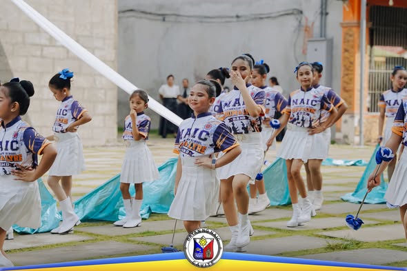 Mga batang babae na nagpeperform ng drill sa paved na plaza, suot ang NAIC na uniporme at may hawak na pom-pom