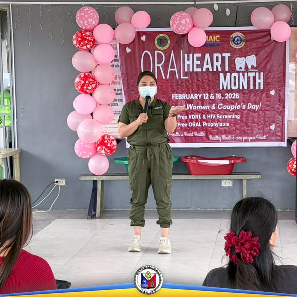 Babaeng nagsasalita sa mikropono sa harap ng banner ng Oral-Heart Month na may pink na lobo sa covered court