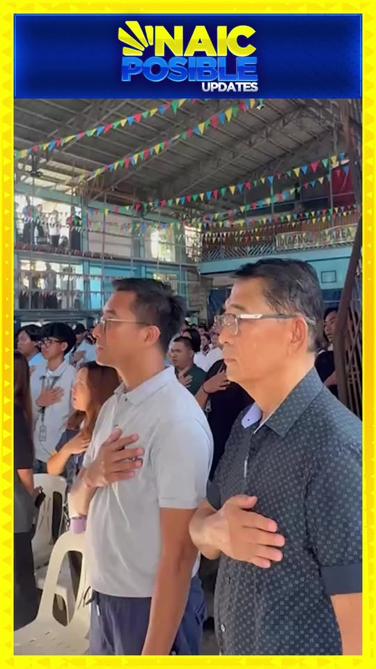 Mga taong nakatayo at nakalagay ang kamay sa dibdib habang nakikinig o umaawit sa loob ng covered court na may bunting.