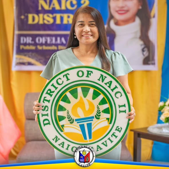 Babaeng nakangiti habang hawak ang sign ng District of Naic II sa harap ng dilaw na kurtina