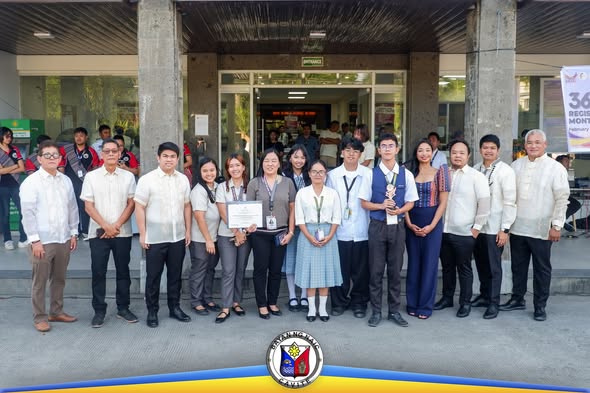 Grupo ng mga kalahok at opisyal na nagpose kasama ang sertipiko at tropeo sa harap ng gusali sa Naic