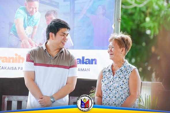 Dalawang tao na nag-uusap at nakangiti sa harap ng banner ng pamamahagi ng school supplies sa Naic