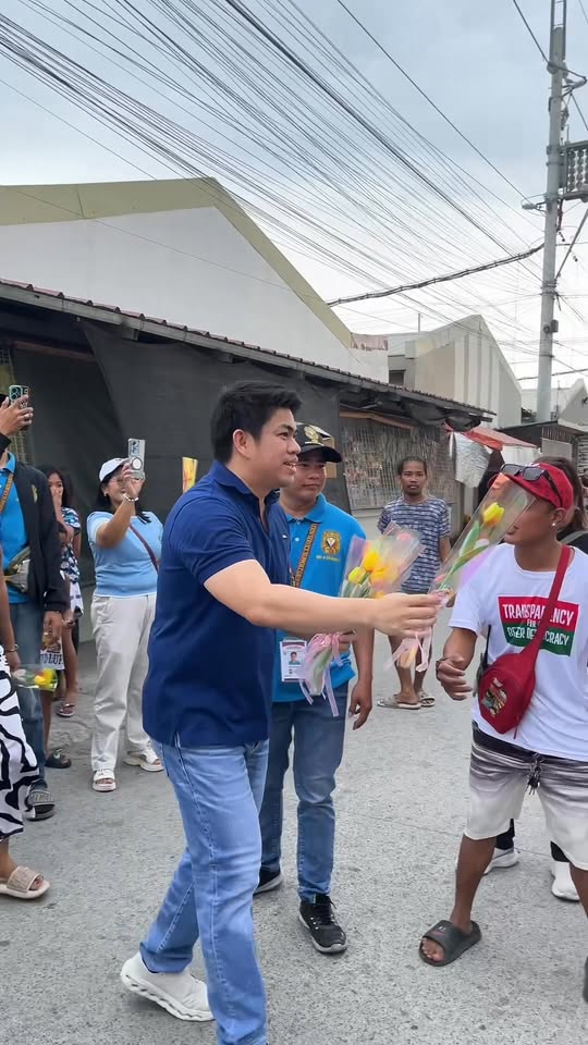 Isang opisyal na namimigay ng bulaklak sa residente sa kalsada ng subdivision habang nanonood ang mga tao
