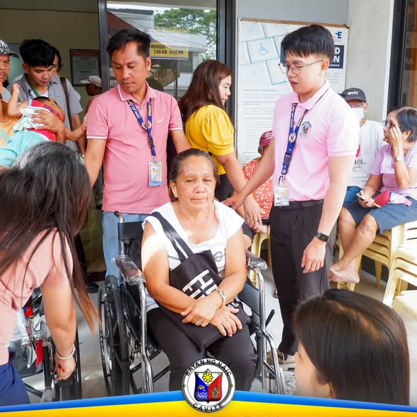 Mga kawani ng Naic habang inaalalayan ang matandang babaeng nakaupo sa wheelchair sa Municipal Hall