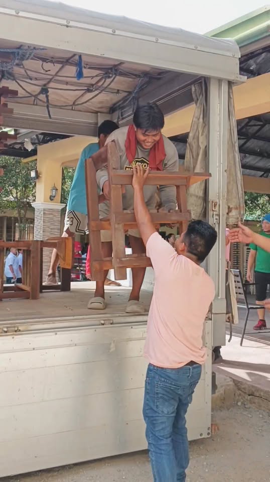 Mga tao naglilipat ng upuang kahoy mula sa truck sa ilalim ng covered area, pamamahagi ng school chairs sa Naic