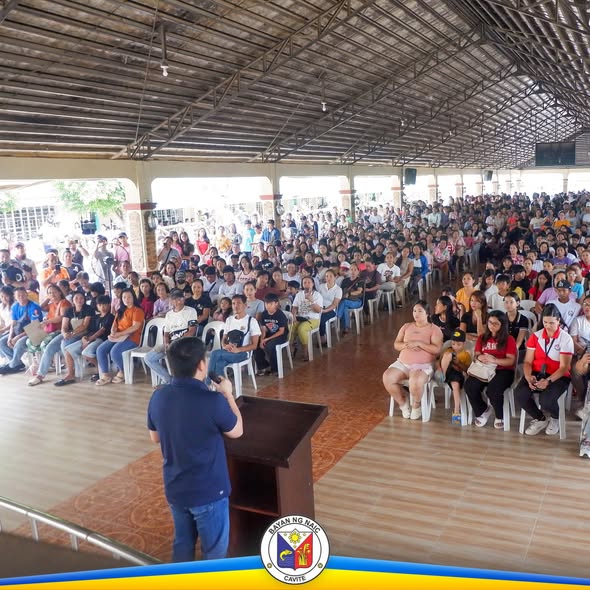 Mga mag-aaral na nakaupo sa covered court habang may nagsasalita sa podium sa pamamahagi ng school supplies