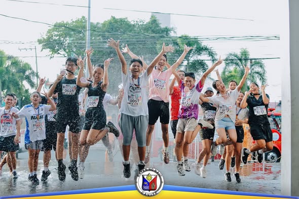 Mga kalahok ng HAKBANG Fun Run 2026 na nagtatalon at nagtataas ng kamay habang nababasa ng tubig sa Naic