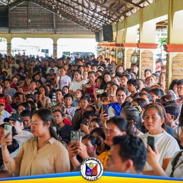 Malaking grupo ng tao sa loob ng covered hall habang may hawak na cellphone, nakikinig at nanonood sa pamamahagi
