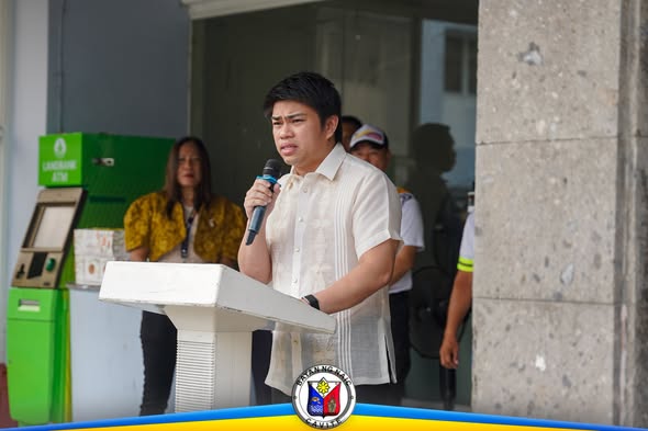 Lalaking nagsasalita sa mikropono sa podium sa flag raising ceremony, may kawani sa likuran