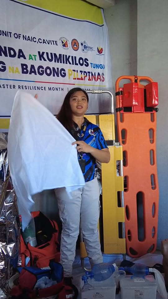 Babaeng nagpapaliwanag habang hawak ang puting tela sa harap ng rescue stretcher at kagamitan ng MDRRMO sa Naic