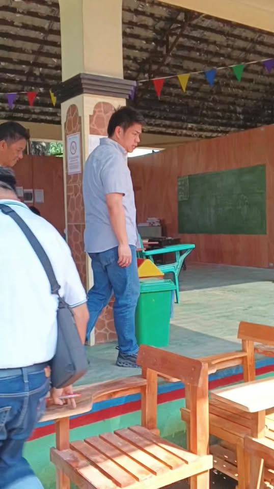 Lalaking nakatayo sa loob ng covered court ng paaralan habang may mga bagong upuang kahoy sa harapan