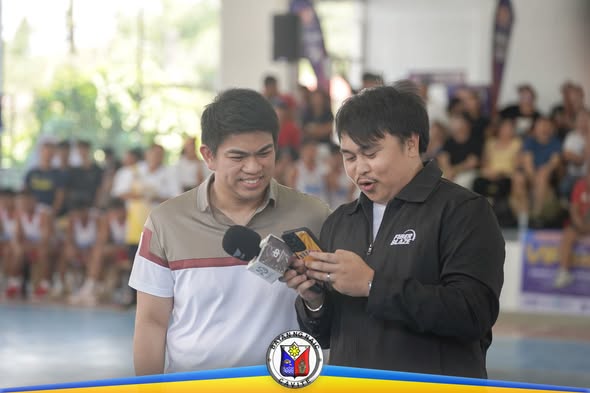 Dalawang lalaki sa loob ng gym habang nagbabalita sa cellphone at mikropono sa Cavite Ballers League