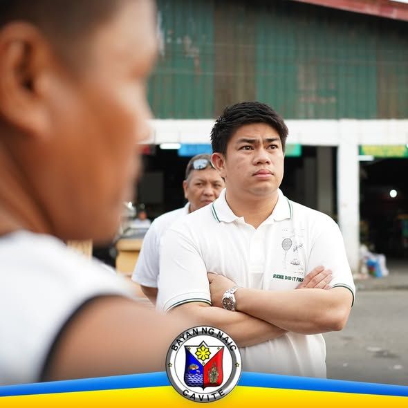 Isang opisyal na nakatayo habang naka-krus ang braso sa palengke, kausap ang mga tao sa harap