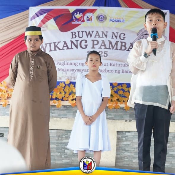 Tatlong bata sa entablado habang nagsasalita ang isa sa Buwan ng Wikang Pambansa 2025 sa Naic