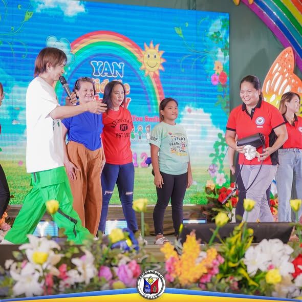 Mga tao sa entablado ng Yan Kiddie Show, may makulay na backdrop ng bahaghari at araw sa Naic