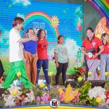 Mga tao sa entablado ng Yan Kiddie Show, may makulay na backdrop ng bahaghari at araw sa Naic