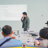 Lalaking nagsasalita sa mikropono sa harap ng screen na may 'Upcoming Activities' sa pulong ng board